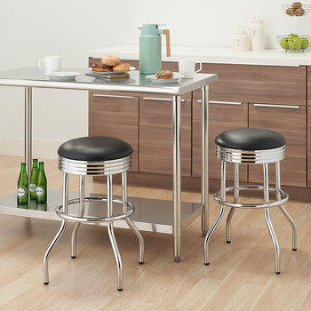 A modern kitchen with a shiny stainless steel counter-height table and two matching bar stools with black padded seats. The table has two mugs, a plate of donuts, and a teal thermos on top, while the lower shelf holds three green bottles.
