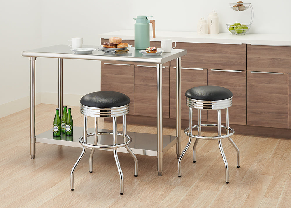 A modern kitchen with a shiny stainless steel counter-height table and two matching bar stools with black padded seats. The table has two mugs, a plate of donuts, and a teal thermos on top, while the lower shelf holds three green bottles.