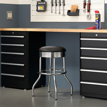A black and silver metal stool sits in front of a long wooden workbench, which is supported by two black metal tool cabinets. Above the bench, tools and a can of WD-40 are organized on a white pegboard.