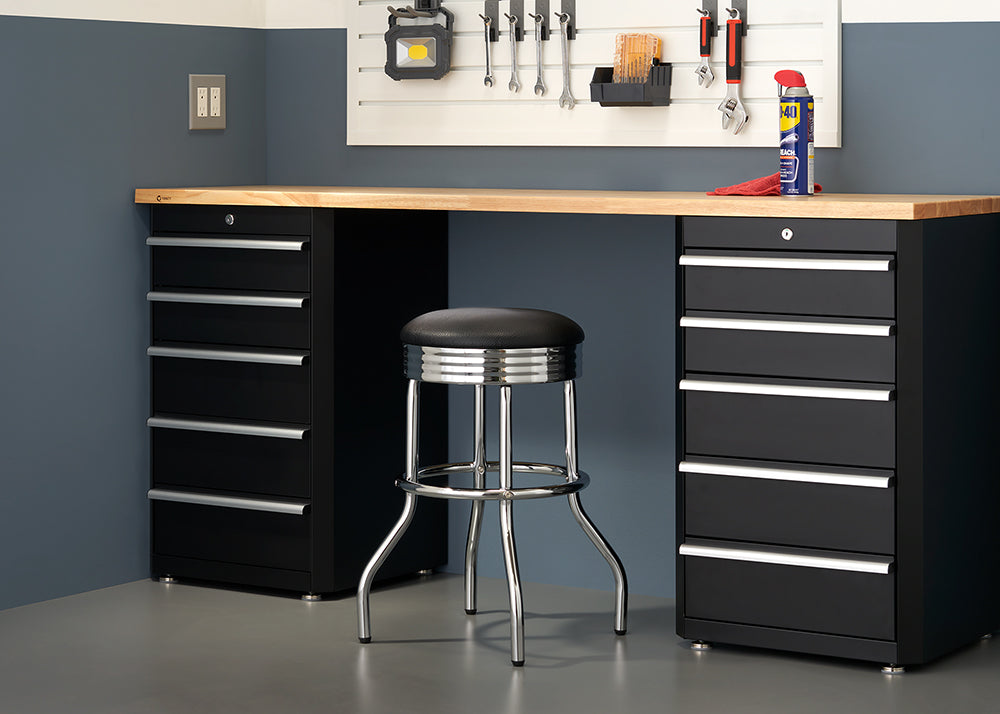 A black and silver metal stool sits in front of a long wooden workbench, which is supported by two black metal tool cabinets. Above the bench, tools and a can of WD-40 are organized on a white pegboard.