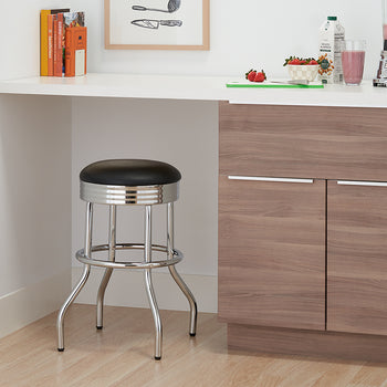 A low-angle shot shows a retro chrome and black bar stool in a modern kitchen. The stool is tucked under a white counter, next to wood-grain cabinets and a blender with a pink smoothie.