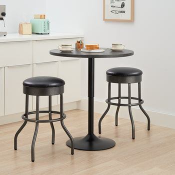 A small, round black bistro table is set with two cups and a plate of toast, situated between two matching black bar stools with black faux leather cushions and curved metal legs. The set sits on a light wood floor in a kitchen area with white cabinets, a silver espresso machine, and a mint green toaster visible in the background.