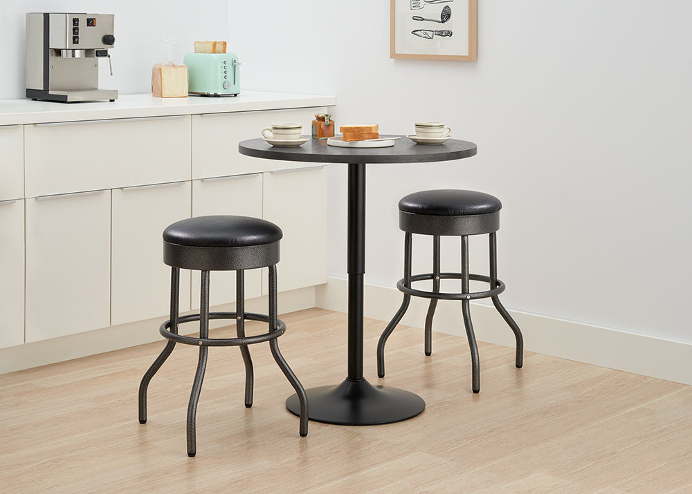 A small, round black bistro table is set with two cups and a plate of toast, situated between two matching black bar stools with black faux leather cushions and curved metal legs. The set sits on a light wood floor in a kitchen area with white cabinets, a silver espresso machine, and a mint green toaster visible in the background.