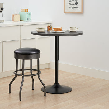 A modern kitchen corner features a small, round black metal bar table and a single matching barstool with a black faux leather seat. On the counter behind the table are a stainless steel espresso machine, a light blue retro toaster, and some bread slices, suggesting a breakfast setting.