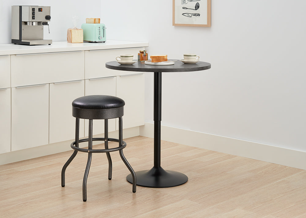 A modern kitchen corner features a small, round black metal bar table and a single matching barstool with a black faux leather seat. On the counter behind the table are a stainless steel espresso machine, a light blue retro toaster, and some bread slices, suggesting a breakfast setting.