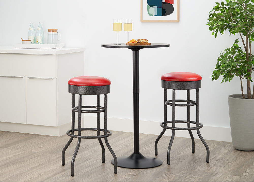 A close-up shot features a round, black pedestal bar table set for two with appetizers and champagne flutes. Two industrial-style bar stools with black metal legs and round red faux-leather seats are tucked under the table in a modern kitchen or bar area.