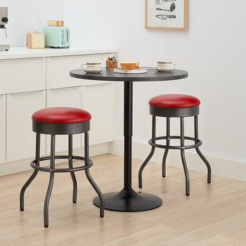 A small, black metal bistro table with a round top stands between two backless bar stools with red faux-leather padded seats. This compact set is placed in a light-colored kitchen with white cabinetry, ready for a quick meal.