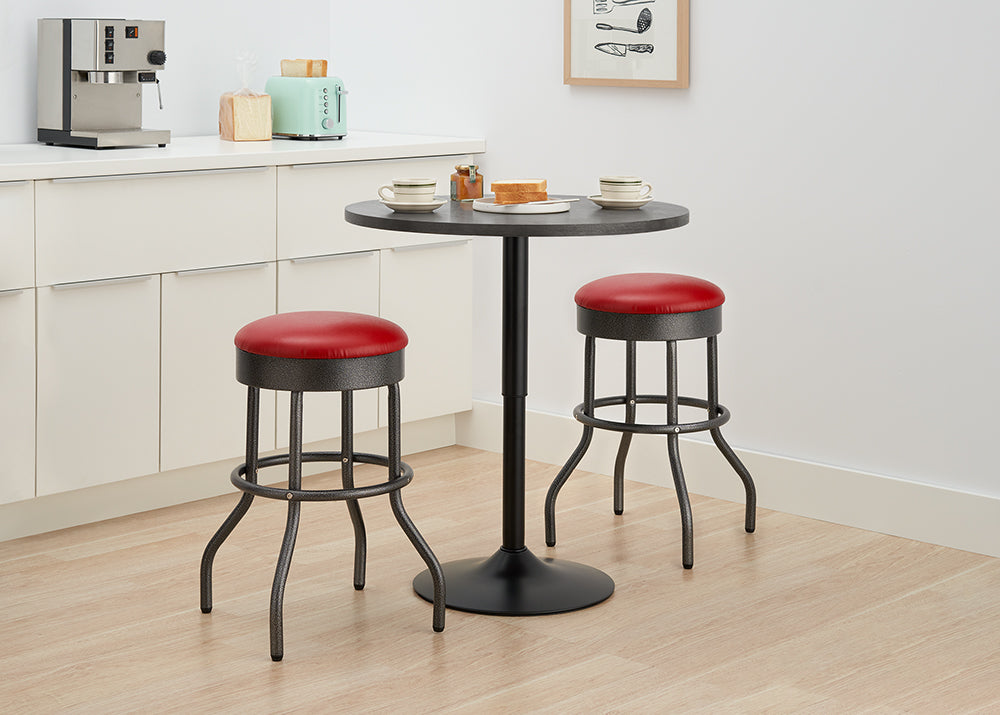 A small, black metal bistro table with a round top stands between two backless bar stools with red faux-leather padded seats. This compact set is placed in a light-colored kitchen with white cabinetry, ready for a quick meal.