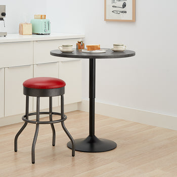 A black, adjustable-height round bar table sits in a kitchen corner, set for two with coffee cups and toast. Beside the table is a retro-style bar stool with a red cushioned seat and a black, wrought-iron base.
