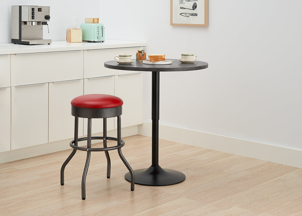 A black, adjustable-height round bar table sits in a kitchen corner, set for two with coffee cups and toast. Beside the table is a retro-style bar stool with a red cushioned seat and a black, wrought-iron base.