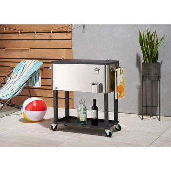 A stainless steel Trinity cooler with a black stand and wheels sits on an outdoor patio. The cooler has a split lid, a bottle opener on the front, and a towel hanging from the side handle.