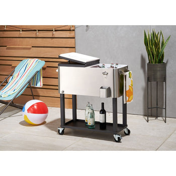 A stainless steel cooler with a partially open white lid sits on a black wheeled stand, filled with ice and various bottled beverages. The cooler has a bottle opener attached to the front and a patterned towel hanging from the side, in an outdoor setting with a beach chair, beach ball, and potted plant visible in the background.