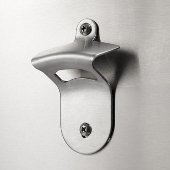 A close-up shot shows a stainless steel, wall-mounted bottle opener affixed to a brushed metal surface with two visible screws. The bottle opener has a classic design with an open mouth for levering bottle caps.