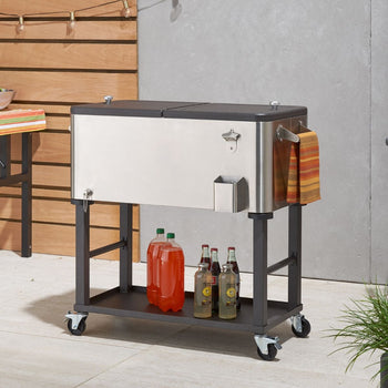 A Trinity 80 Quart Stainless Steel Detachable Cooler on a wheeled stand with a bottom shelf. Several bottles and a towel are placed on or beside the cooler, suggesting it's being used for outdoor entertaining.