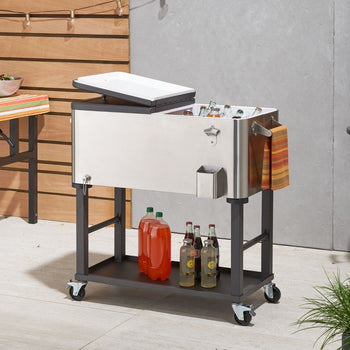 A stainless steel drinks cooler trolley on wheels sits on a patio. The cooler is partially open, revealing ice and various bottled and canned beverages inside; additional bottled drinks are on the shelf below.