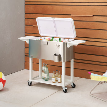 A large, stainless steel outdoor cooler with its lid open, revealing bottles and cans inside, stands on a white wheeled cart. The cooler is positioned on a paved patio next to a wooden slatted wall, with a blue inflatable tube, a beach ball, and a striped beach chair visible nearby.
