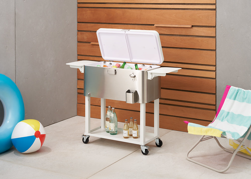 A large, stainless steel outdoor cooler with its lid open, revealing bottles and cans inside, stands on a white wheeled cart. The cooler is positioned on a paved patio next to a wooden slatted wall, with a blue inflatable tube, a beach ball, and a striped beach chair visible nearby.