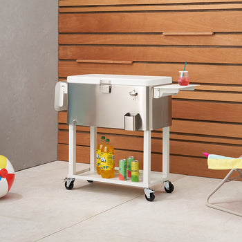 
A sleek, stainless steel outdoor cooler on wheels stands beside a wooden wall, with a colorful beach ball, a blue inflatable ring, and a striped beach chair nearby. The cooler has a lower shelf holding drinks and features a bottle opener and side tray.