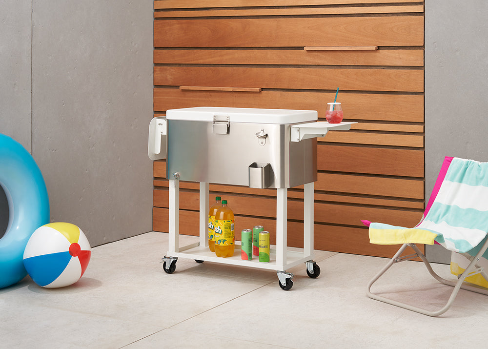 
A sleek, stainless steel outdoor cooler on wheels stands beside a wooden wall, with a colorful beach ball, a blue inflatable ring, and a striped beach chair nearby. The cooler has a lower shelf holding drinks and features a bottle opener and side tray.