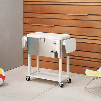 A polished stainless steel cooler on a white rolling stand is positioned on a tiled patio. To the left are a blue inflatable and a colorful beach ball, while a yellow and pink striped beach chair is visible on the right.
