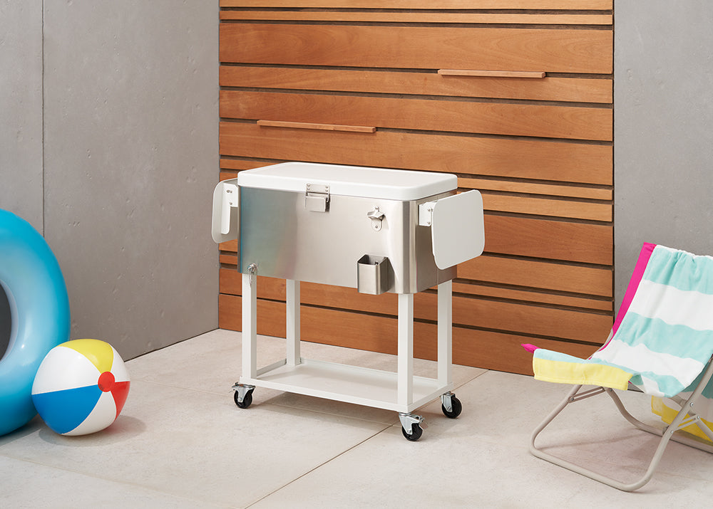A polished stainless steel cooler on a white rolling stand is positioned on a tiled patio. To the left are a blue inflatable and a colorful beach ball, while a yellow and pink striped beach chair is visible on the right.