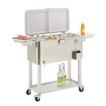 A stainless steel outdoor cooler on wheels with its lid open, revealing a variety of drinks on ice. The cooler has two side tables, one holding a plate of watermelon slices and the other an orange soda bottle, with two more bottled drinks on a shelf below.