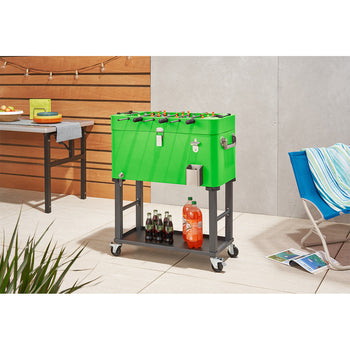 A bright green cooler shaped like a foosball table stands on a wheeled gray metal cart on a patio. The cooler has foosball rods with players on top, a bottle opener, and a catch bin, while the cart's lower shelf holds several bottles of soda.