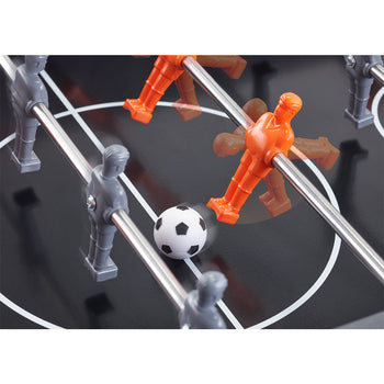 A close-up shot shows a foosball table in action. A black and white soccer ball is positioned between gray and orange player figures attached to metal rods, suggesting a game in progress.