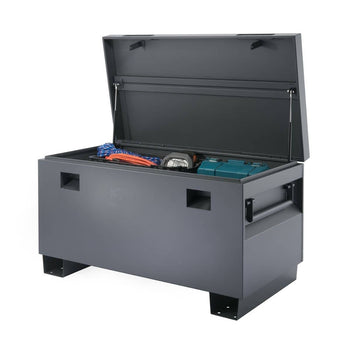 A dark gray, heavy-duty Trinity job site box is shown slightly open, revealing some tools and equipment inside. The sturdy metal box has handles on the sides and rests on a raised base.