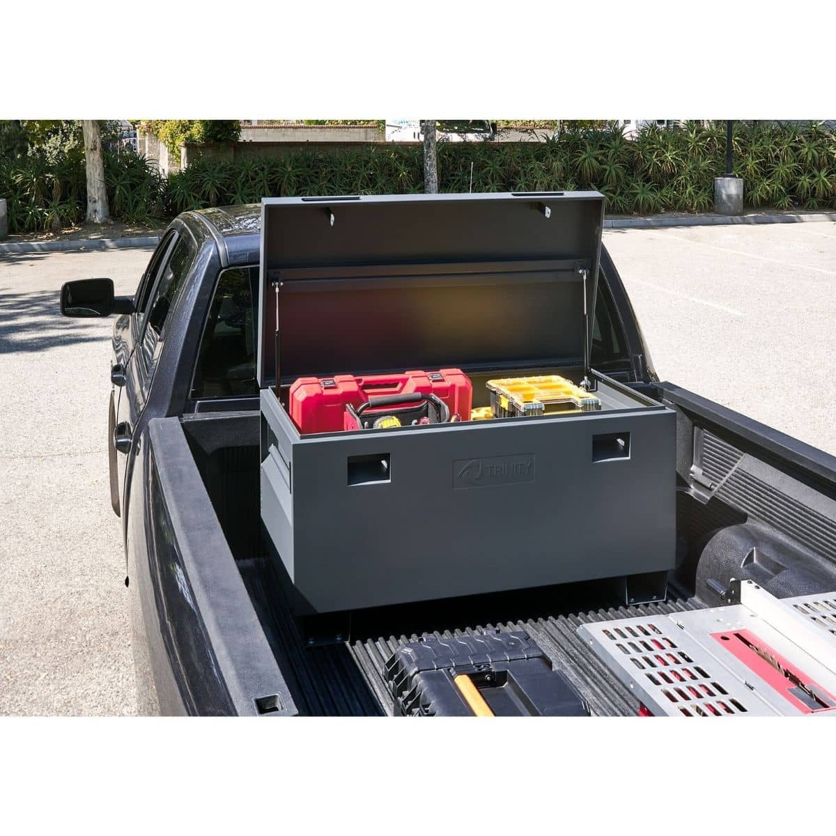A dark gray Trinity job site box with its lid open sits in the bed of a black pickup truck. Inside the box are red and yellow toolboxes, and the truck bed also contains other tools and equipment.
