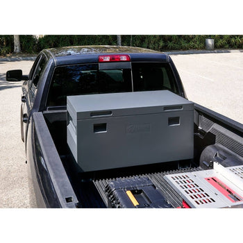 A gray Trinity job site box is positioned in the bed of a black pickup truck. Other tools and equipment are partially visible around the toolbox in the truck bed.