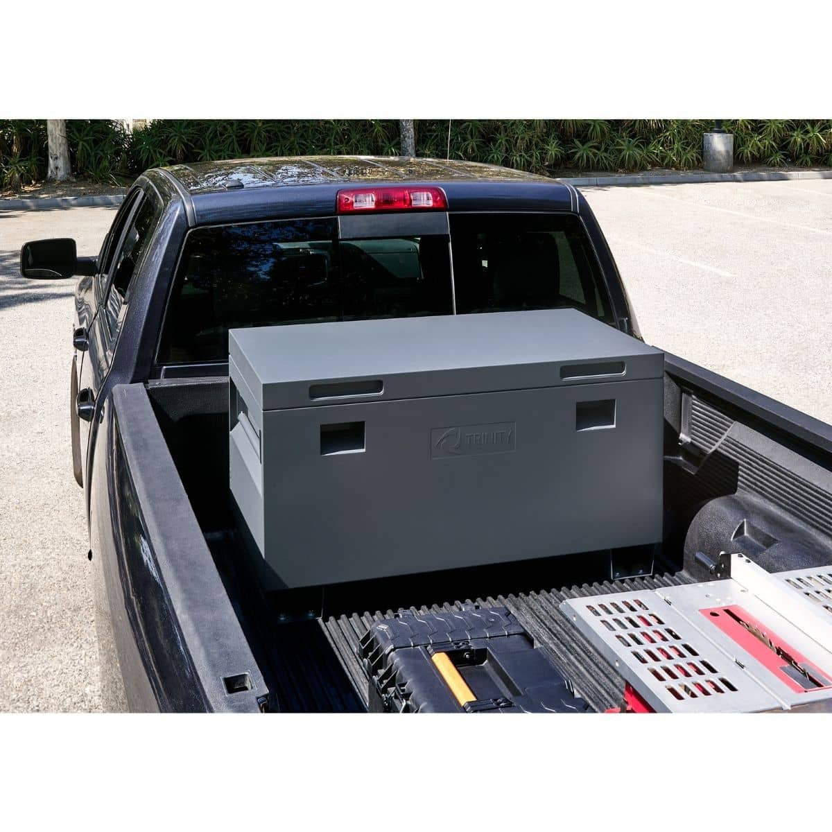 A gray Trinity job site box is positioned in the bed of a black pickup truck. Other tools and equipment are partially visible around the toolbox in the truck bed.