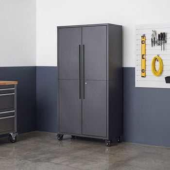 A tall, closed dark gray steel storage cabinet featuring long vertical handles and caster wheels stands in a garage setting. It is set against a blue and white wall alongside a tool organizer slatboard and a rolling metal workbench.