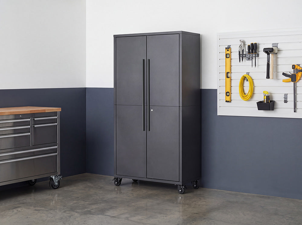 A tall, closed dark gray steel storage cabinet featuring long vertical handles and caster wheels stands in a garage setting. It is set against a blue and white wall alongside a tool organizer slatboard and a rolling metal workbench.