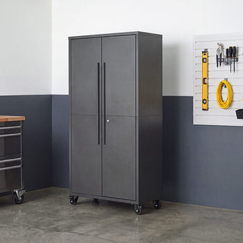 A tall, closed dark gray steel storage cabinet with vertical door handles stands on caster wheels in a workshop setting. The mobile cabinet is positioned next to a stainless steel workbench and a wall slatboard mounted with organized tools.