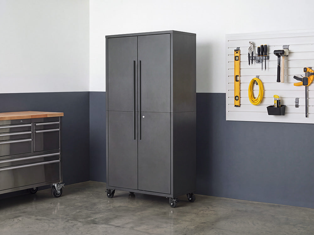 A tall, closed dark gray steel storage cabinet with vertical door handles stands on caster wheels in a workshop setting. The mobile cabinet is positioned next to a stainless steel workbench and a wall slatboard mounted with organized tools.