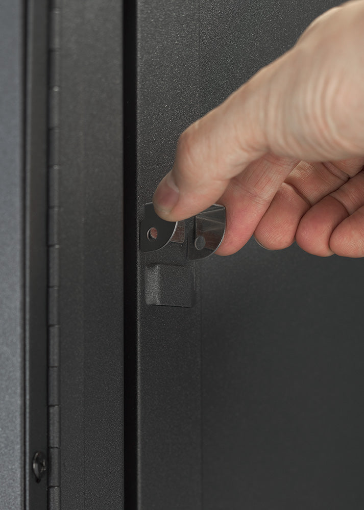 A close-up view shows a person's hand operating the internal metal latch mechanism on the door edge of a dark gray steel storage cabinet. The detail highlights the functional door lock latch assembly on the powder-coated cabinet door.