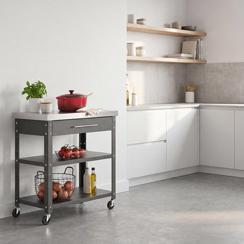 A dark gray rolling kitchen island with a stainless steel top stands against a plain white wall in a modern, sunlit kitchen. The cart is styled with a potted plant and a red Dutch oven on top, fresh tomatoes and onions on its lower shelves, and sits next to white minimalist cabinetry and open wooden shelving.