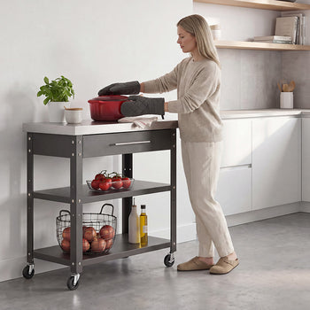 A blonde woman wearing a neutral sweater, matching trousers, and oven mitts adjusts a red Dutch oven on a dark gray rolling kitchen cart. The cart features a stainless steel countertop, a single top drawer, and open shelves holding fresh produce and cooking oils in a bright, modern kitchen setting.