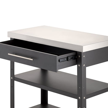 A close-up view shows the upper portion of a dark gray metal kitchen cart with a stainless steel countertop against a white background. Its top drawer is pulled half open on smooth metal glides, revealing an empty interior and two lower open shelves beneath.