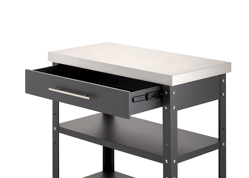 A close-up view shows the upper portion of a dark gray metal kitchen cart with a stainless steel countertop against a white background. Its top drawer is pulled half open on smooth metal glides, revealing an empty interior and two lower open shelves beneath.