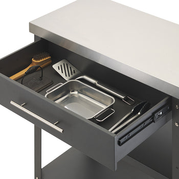 A close-up view shows the open drawer of a dark gray kitchen cart topped with a stainless steel surface. Inside the drawer, several cooking and grilling tools are neatly organized, including a metal spatula, tongs, a small roasting pan, a cleaning brush, and a press.