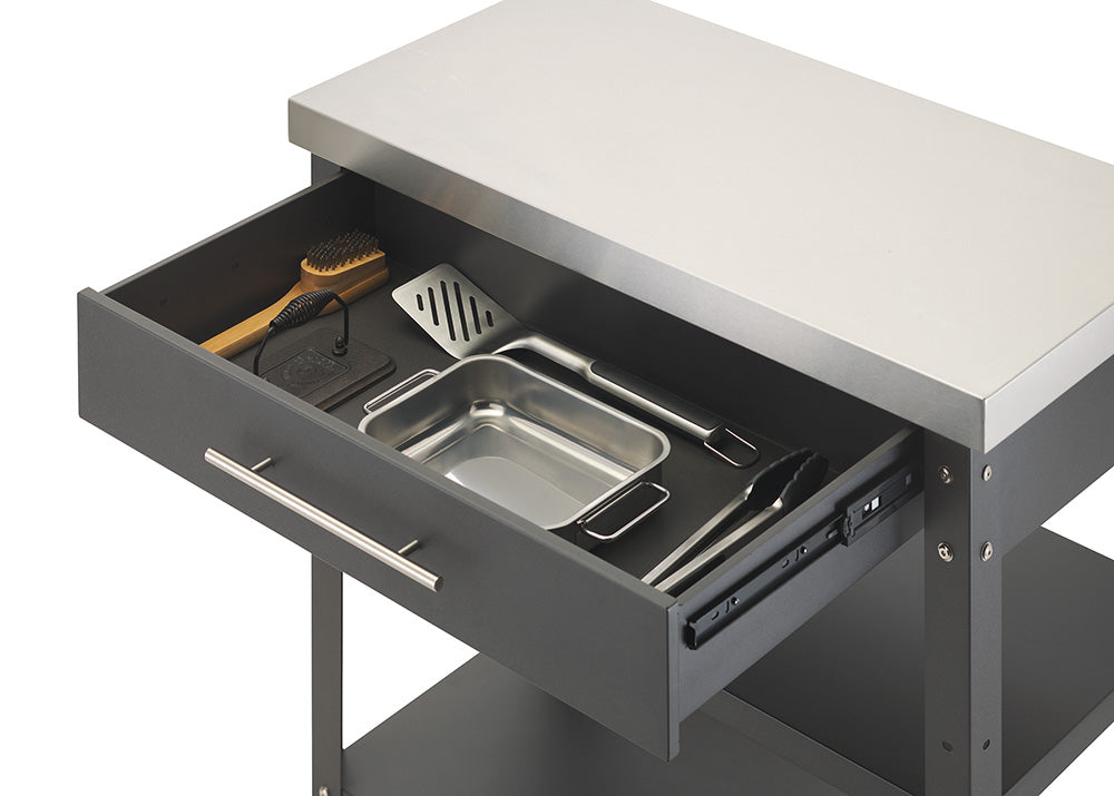 A close-up view shows the open drawer of a dark gray kitchen cart topped with a stainless steel surface. Inside the drawer, several cooking and grilling tools are neatly organized, including a metal spatula, tongs, a small roasting pan, a cleaning brush, and a press.