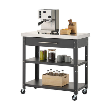 A black rolling kitchen cart features a stainless steel countertop, a single top drawer, and two lower open shelves against a plain white background. On top sits an espresso machine alongside wooden organizer caddies, while the middle shelf holds mugs, coffee beans, and tea tins, and the bottom shelf holds a wooden crate filled with glass bottles.