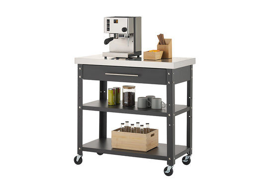 A black rolling kitchen cart features a stainless steel countertop, a single top drawer, and two lower open shelves against a plain white background. On top sits an espresso machine alongside wooden organizer caddies, while the middle shelf holds mugs, coffee beans, and tea tins, and the bottom shelf holds a wooden crate filled with glass bottles.