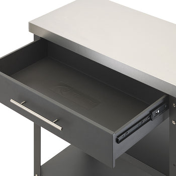 An angled, close-up shot displays the top drawer of a dark gray kitchen cart pulled fully open beneath a stainless steel countertop. The interior of the drawer is completely empty, lined with a black mat that features an embossed Trinity logo in the center.