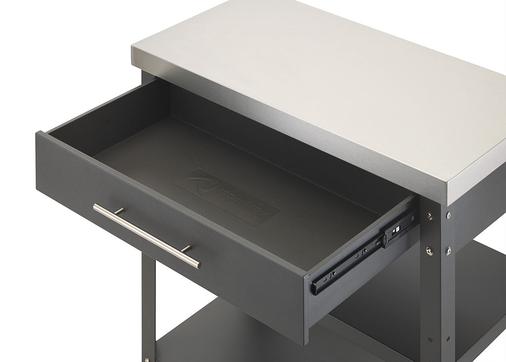 An angled, close-up shot displays the top drawer of a dark gray kitchen cart pulled fully open beneath a stainless steel countertop. The interior of the drawer is completely empty, lined with a black mat that features an embossed Trinity logo in the center.