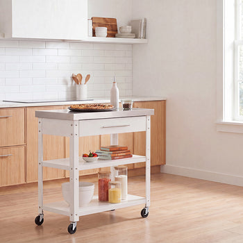 A white rolling kitchen island with a stainless steel countertop stands in the center of a bright, wooden-accented kitchen. It holds a baked tart and oil bottle on top, while its lower shelves are styled with cookbooks, small bowls, and storage jars filled with ingredients.