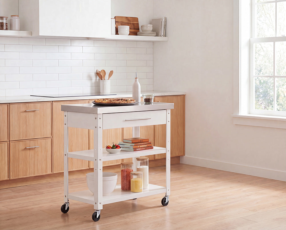 A white rolling kitchen island with a stainless steel countertop stands in the center of a bright, wooden-accented kitchen. It holds a baked tart and oil bottle on top, while its lower shelves are styled with cookbooks, small bowls, and storage jars filled with ingredients.
