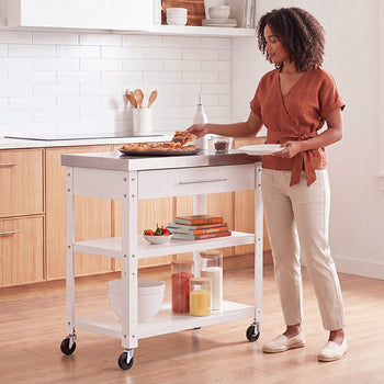 A woman with curly hair, wearing a terracotta wrap top and beige trousers, serves a slice of tart from a white rolling kitchen island. The cart features a stainless steel top, a drawer, and lower open shelves styled with cookware and cookbooks in a bright, modern kitchen.